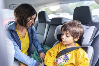 Smiling mother fastening a seat belt for her son in a child safety seat inside a car, ensuring his