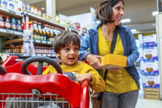 Mother holding a box of cookies next to her son sitting in a shopping cart, enjoying their grocery