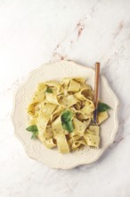 Fresh papardelle pasta, with basil and pesto sauce, is served, on a white plate, with a fork held
