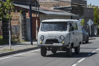 White pickup truck drives past old buildings on a quiet street, UAZ Buchanka, Armenia