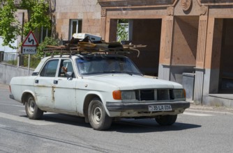A white car with roof luggage drives through an urban environment with old buildings, GAZ Volga,