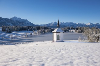 Snowy chapel with mountain backdrop and clear sky, surrounded by forests, Hegratsrieder See, near