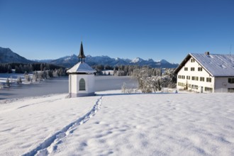 Snowy landscape with chapel and farmhouse against mountain backdrop under blue sky, Hegratsrieder