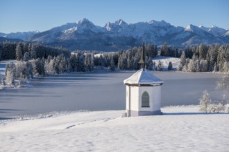 Chapel on the shore of a frozen lake surrounded by snow and mountains under a blue sky,