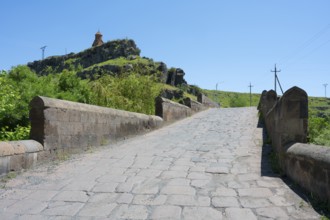 A stone bridge leads to a hill with a church under clear sky and green landscape, St. Sarkis church