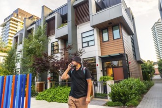 Young man with backpack is using his phone while walking in front of modern townhouses in