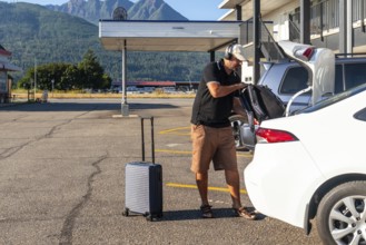 Tourist wearing headphones is loading luggage into car trunk in motel parking lot, preparing for
