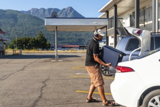 Tourist wearing headphones is loading a suitcase into the trunk of his car in the parking lot of a