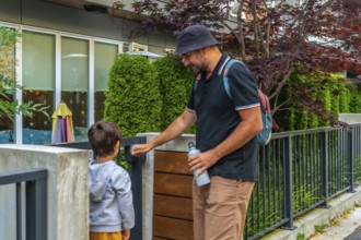 Father holding a reusable water bottle and wearing a bucket hat opens the gate for his son at a