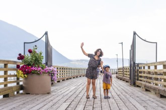 Cheerful mother and her young son are waving while standing on a picturesque wooden pier in salmon