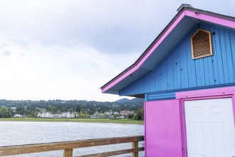 Pink and blue painted building standing on a wooden pier overlooking shuswap lake and the cityscape