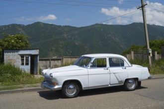 White vintage car on a country road with mountain backdrop and blue sky in summer, GAZ M-21 Volga,