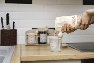 A person is pouring sugar into a container in a kitchen. The countertop is made of wood and has