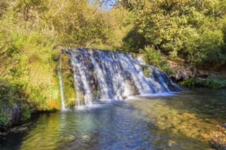 A small waterfall flows over green plants into a clear river under a sunny sky, El Kelaa torrent in