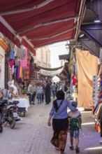 Covered bazaar with colorful fabrics and people, a woman with a child walks through the market