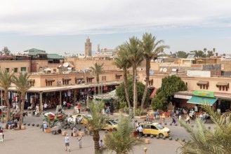 Bustling market scene with palm trees and buildings, a minaret and a blue sky in the background