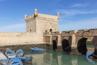 Old stone bridge with small colorful boats in a quiet harbor and a historic fortress in the