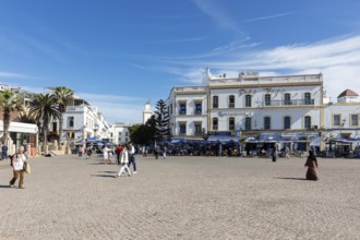 People walk on a wide square surrounded by historic buildings in clear skies