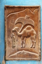 Wood carving of a camel depicting a person in traditional clothing against a hill backdrop,