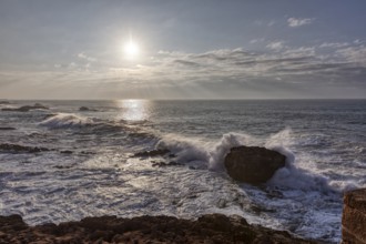 Waves crash against rocks in the ocean at sunset under a cloudy sky