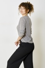 A model with curly hair strikes a pose in a casual grey sweater and black pants. The background is
