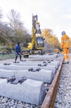 Concrete ties are laid with an excavator along a railway line, supervised by workers, track