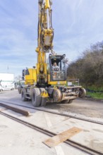 A yellow excavator on a construction site with tracks in the foreground. The sky is clear and the