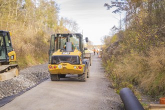 A yellow excavator is standing on a construction site, surrounded by autumn trees. Workers can be