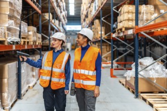 Two women wearing hard hats and safety vests are analyzing inventory by scanning product barcodes