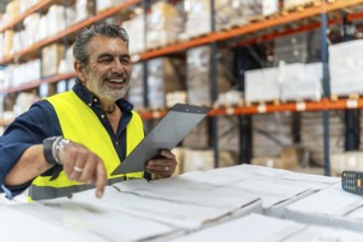 Male logistics supervisor wearing safety vest pointing and holding clipboard, smiling while
