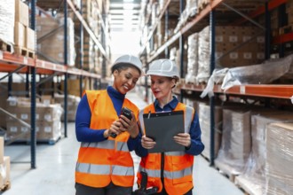 Two women warehouse workers in safety hats and vests are collaborating, scanning inventory with a