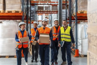 Diverse team of smiling warehouse workers in hard hats and vests carrying boxes and tools through a