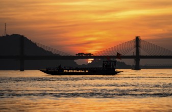 A boat sails on the river Brahmaputra during sunset, with the silhouette of a cable-stayed bridge