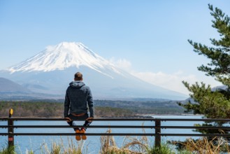 Young man sitting on railings next to a road and looking across the lake to Mt Fuji volcano, Motosu