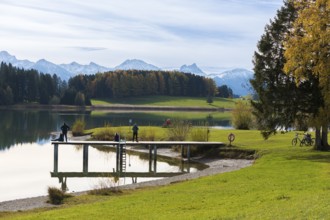 People walk along the jetty with views of Lake Forggensee and surrounding mountains, autumn trees