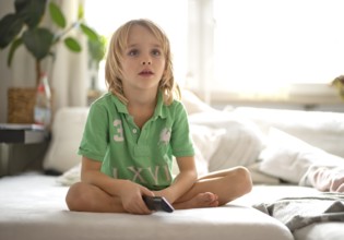 Child, boy, 7 years old, blonde, sitting on sofa, with remote control in hand, watching TV,