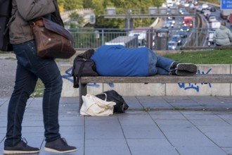 Man sleeping on a bench, travelling bag serves as pillow, in the city centre, passers-by walk by