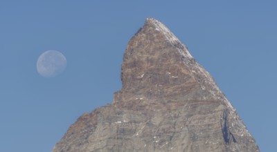 The Matterhorn mountain peak rises steeply against a bright blue sky. The full moon glistens gently