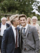 A male couple in a suit, one kissing the other on the cheek, surrounded by guests outdoors,