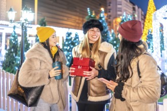 Three cheerful young women holding christmas gifts and hot drinks, enjoying a winter evening in a