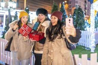 Three cheerful female friends holding christmas gifts and taking a selfie at a decorated outdoor