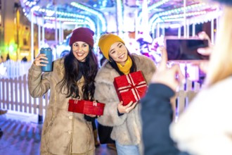 Two happy women holding christmas gifts posing for a picture in front of a carousel at a christmas