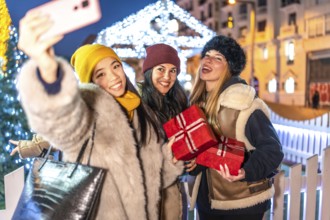 Three cheerful women holding christmas gifts taking a selfie with smartphone at christmas market in