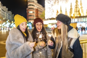 Three happy young women holding burning sparklers, enjoying christmas night in a brightly