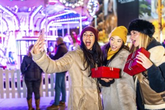 Three cheerful women holding christmas gifts taking a selfie in front of a carousel at night in a