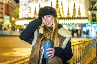 Stylish woman holding reusable cup with hot drink, enjoying magical christmas lights and festive