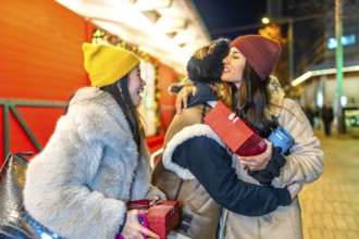 Three female friends exchanging christmas presents and hugging at a christmas market at night