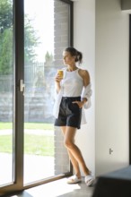A woman dressed in casual summer attire stands by large windows, sipping a smoothies. Natural light
