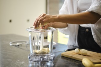 Sliced bananas are placed in a blender while a sunny kitchen provides a bright backdrop. Fresh