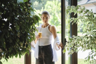 A young woman stands at the entrance of a bright garden space, holding a glass of iced beverage.